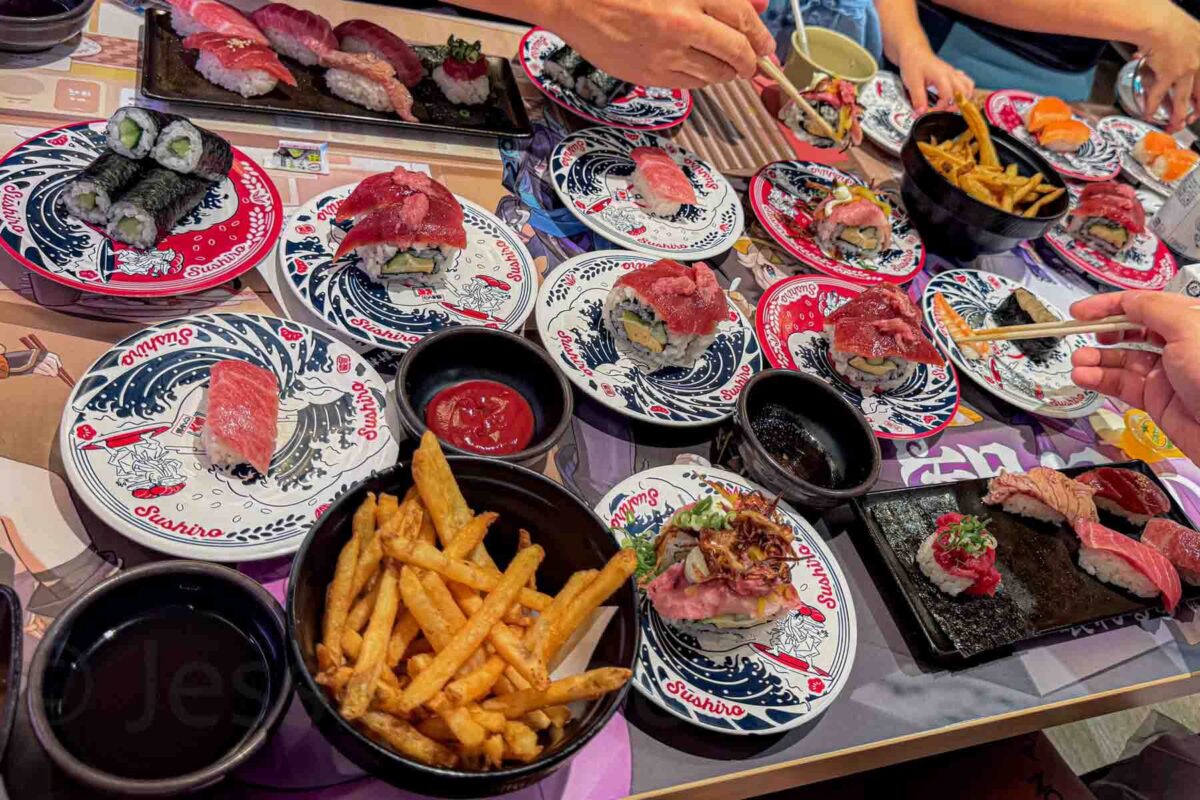 Colorful plates of sushi, tuna, french fries, and dipping sauces spread across a busy table with multiple hands and chopsticks reaching for food at Sushiro in Osaka. A casual setting where kids can try different foods in Japan.