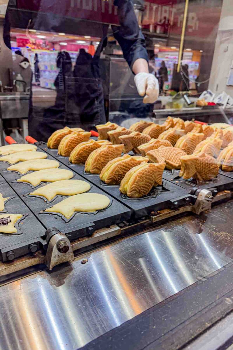 Fresh fish shaped pastries, taiyaki, cooking on a hot griddle while a vendor prepares them behind a glass counter. The warm street snack showcases a fun and kid friendly treat in Japan.