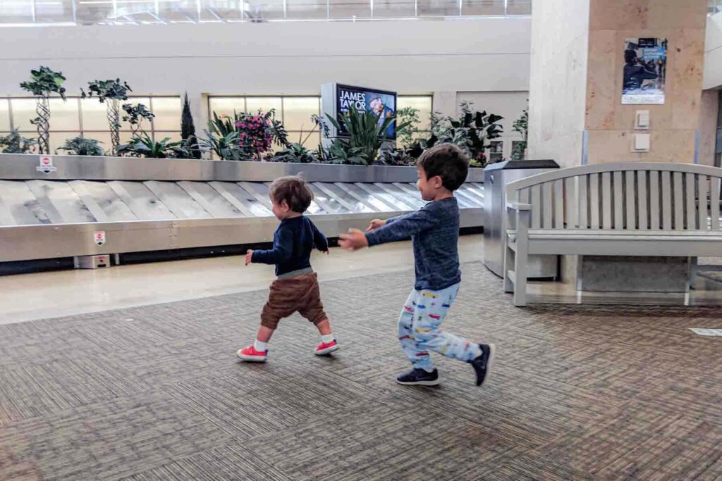 Two young toddler brothers run and play near an airport baggage claim carousel while waiting for luggage. The open space allows them to burn energy after a flight. This stage can feel like the hardest age to travel with a child when toddlers want to move constantly.