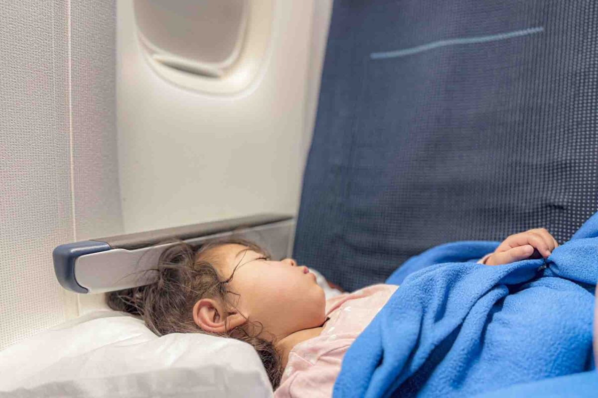 Toddler sleeps peacefully in an airplane seat with head on a pillow and covered by a blue blanket showing how to survive a long flight with kids.