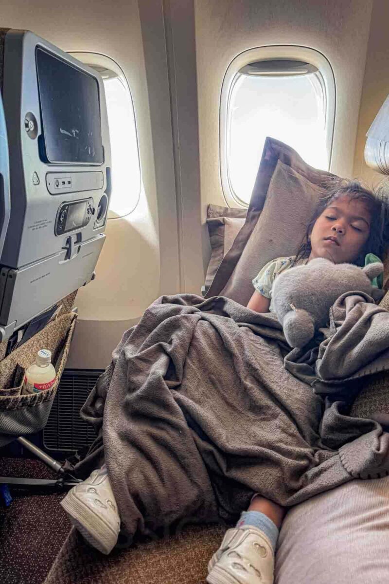 Child sleeps comfortably in a window seat propped up with a pillow and under a blanket holding a stuffed toy during a long haul flight with a toddler from Japan.