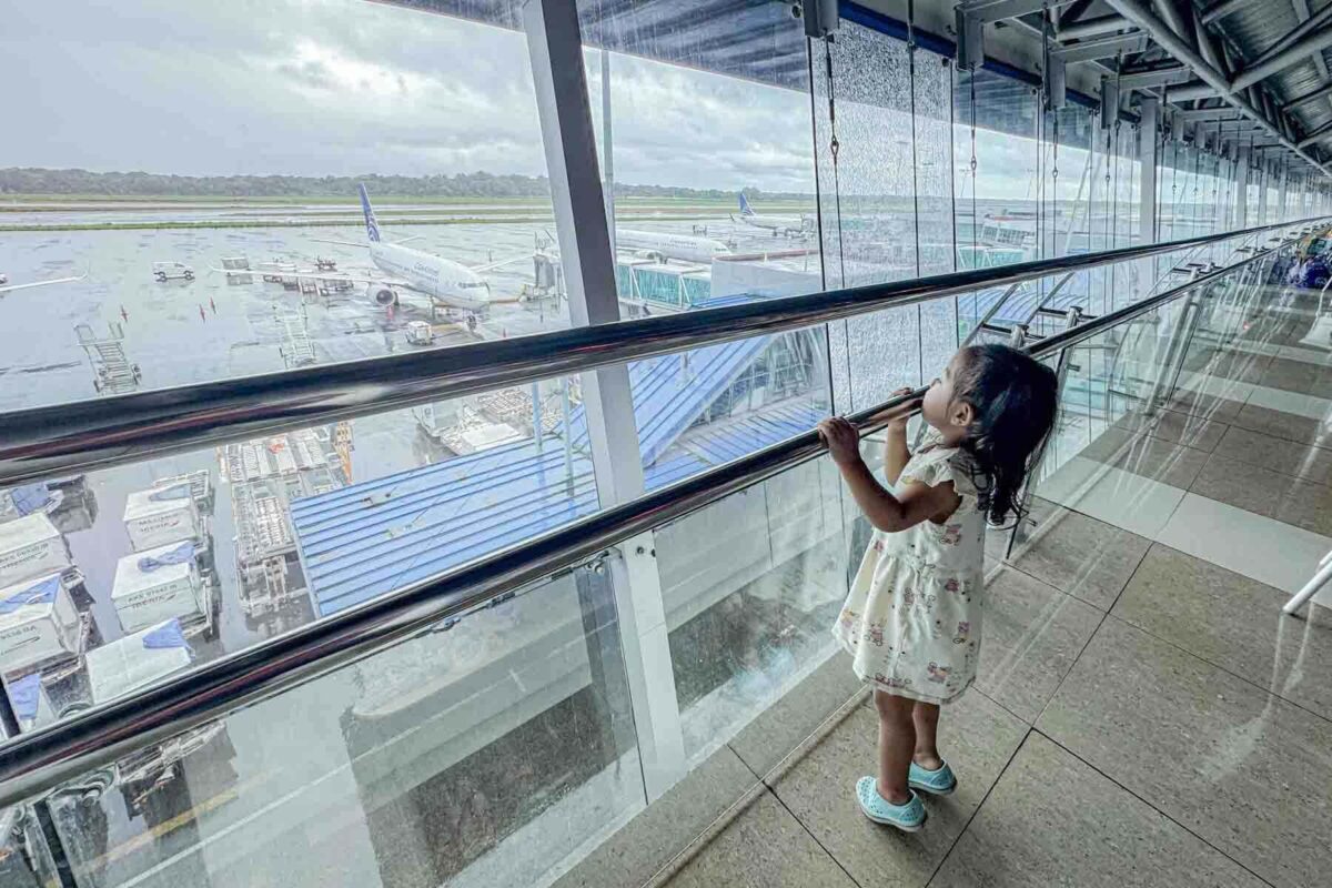 Toddler looks out through airport glass at airplanes parked on the runway on a cloudy rainy day before flying long distance from Panama City.