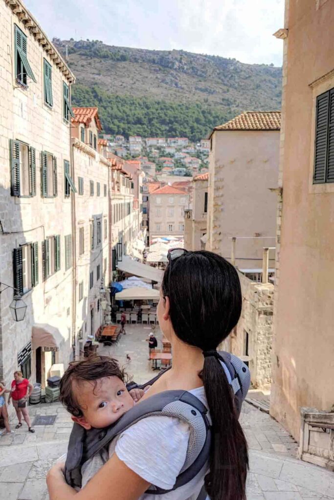 Baby peeks out from a carrier as mother looks over a stone lined street in Dubrovnik, Croatia. Historic buildings and hillside views stretch into the distance.