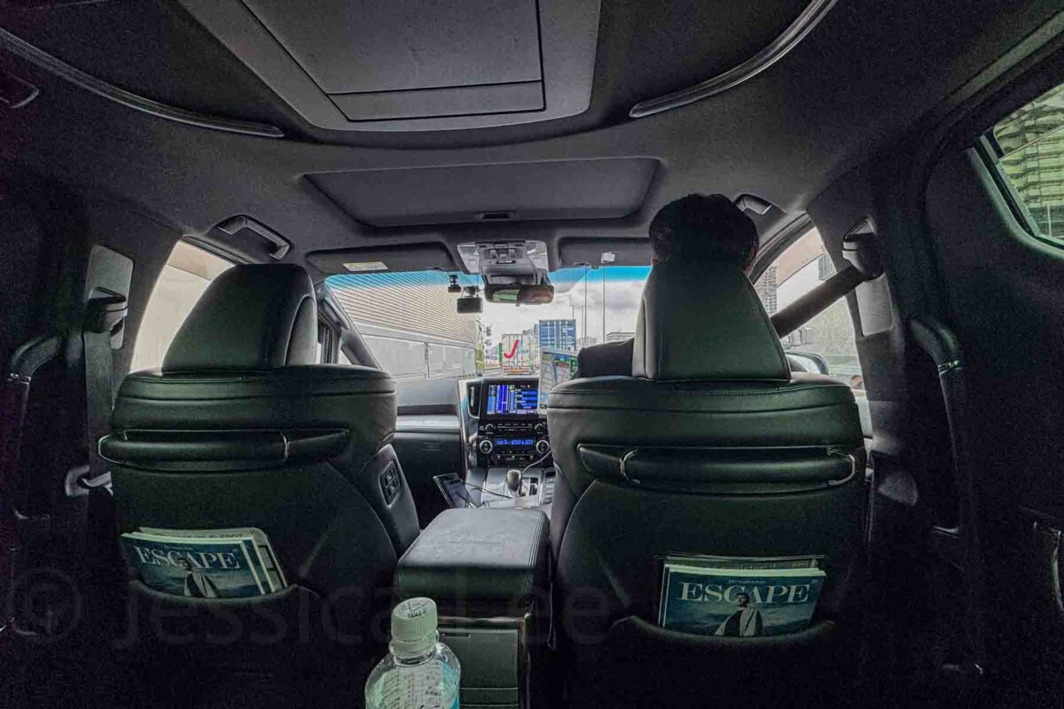 Interior view of a spacious van with black leather seats and a driver visible through the windshield. Presents a private transfer option for comfortable family transportation in Japan.