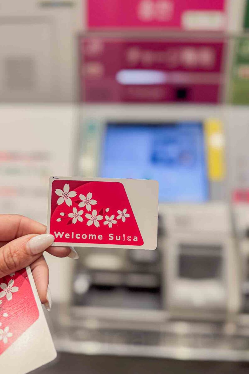 Hand holds a red and white card labeled "Welcome Suica" with a cherry blossom design in front of a ticket machine. Shows an IC prepaid transit card used for trains buses and payments across Japan.