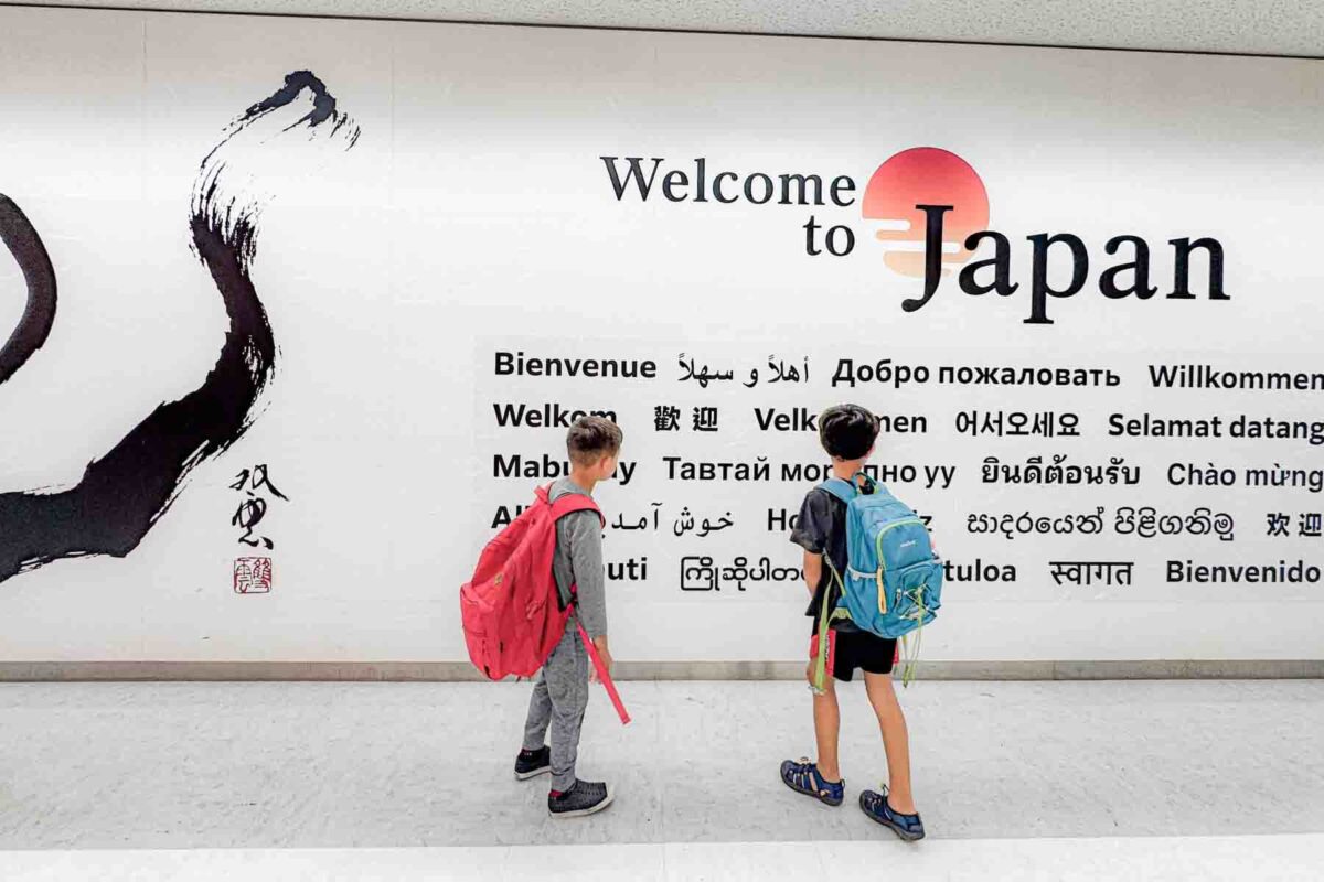 Two kids with backpacks stand in front of a large airport wall sign at Narita airport outside Tokyo that reads "Welcome to Japan" surrounded by greetings in multiple languages. The scene captures the excitement of arriving in Japan with kids as they pause to look at the welcome display.