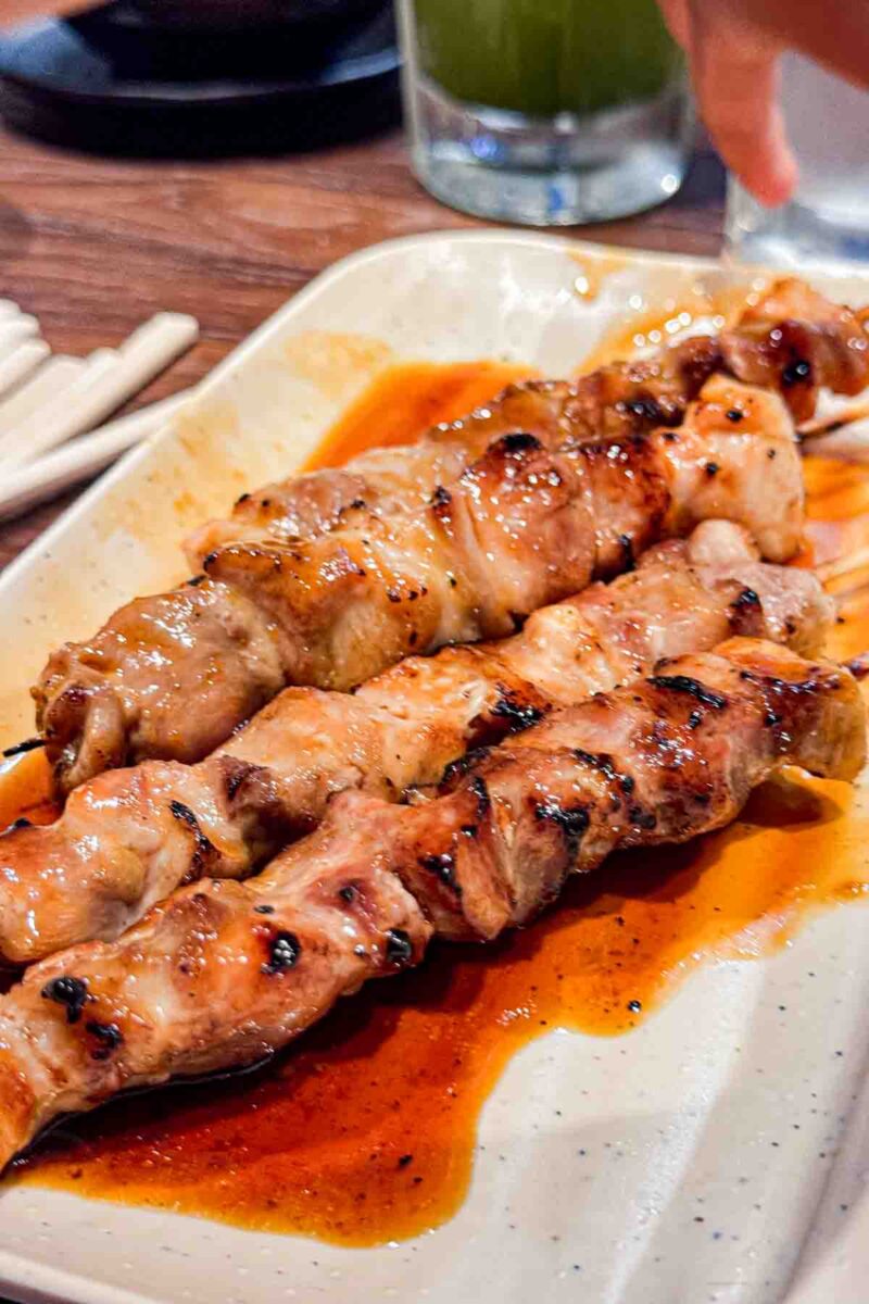 Close up of grilled chicken yakitori skewers coated in a glossy soy sauce on a ceramic plate with chopsticks nearby. 