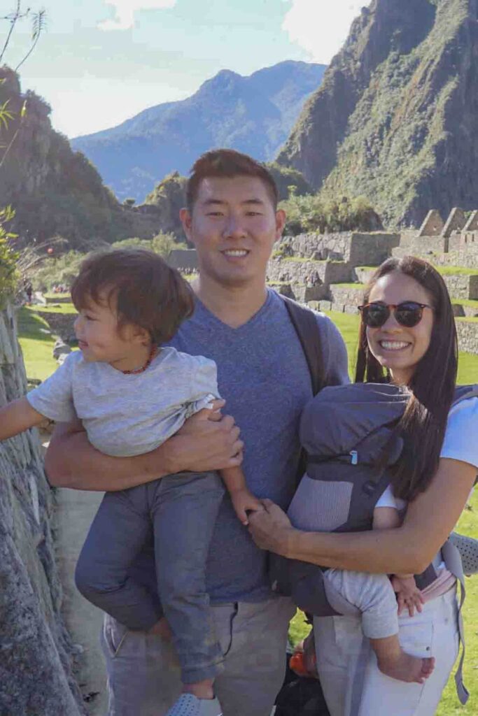 Young family stands together at Machu Picchu in Peru with one parent holding a toddler trying to pull away and the other wearing a sleeping baby in a carrier. The dramatic green mountains and stone ruins rise behind them. This image demonstrates the contrast and differences between traveling with a baby vs toddler.