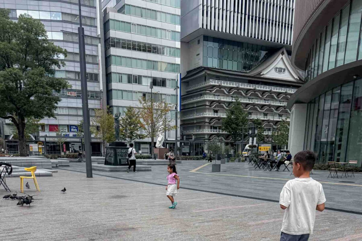 Two children playing in an open city plaza in Namba, Osaka in Japan surrounded by modern buildings and pedestrians. Captures the freedom of exploring on foot when getting around Japan with kids.