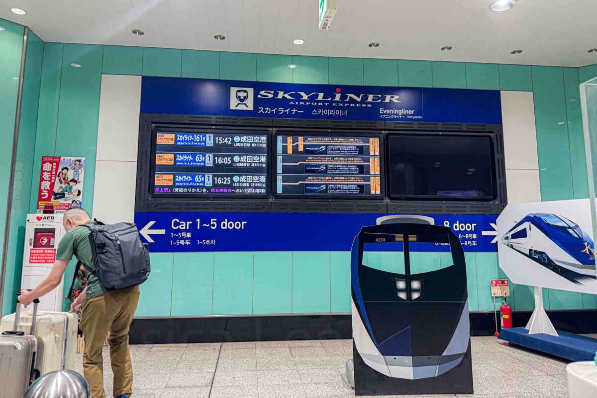 A large digital board displays Skyliner airport express train schedule and times with signage reading "Car 1-5 door" and "8 door" inside a Tokyo station. Clear schedules like this help families plan Tokyo transportation with kids when traveling to and from the airport.