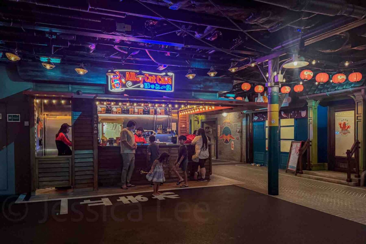 A family sits at the themed "Alley Cat Cafe" counter in the San Fransokyo area, with neon signage and hanging lanterns creating a lively street market atmosphere. This scene highlights immersive dining and snack options onboard the Disney Adventure cruise.