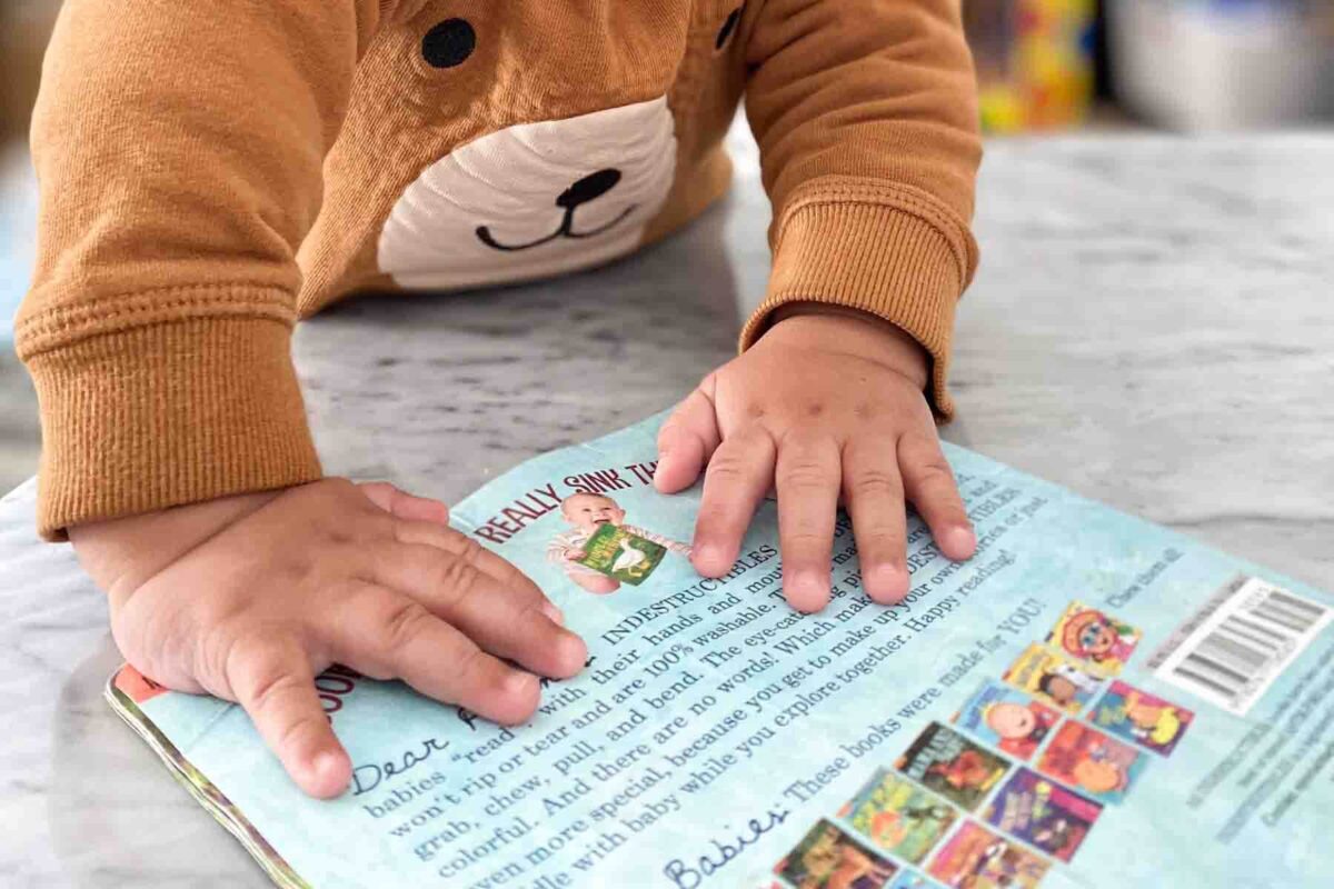 Baby’s hands exploring the back of a soft “Indestructibles” book with visible text including “Really Silly Animals” and care instructions, a durable baby travel toy for flights.