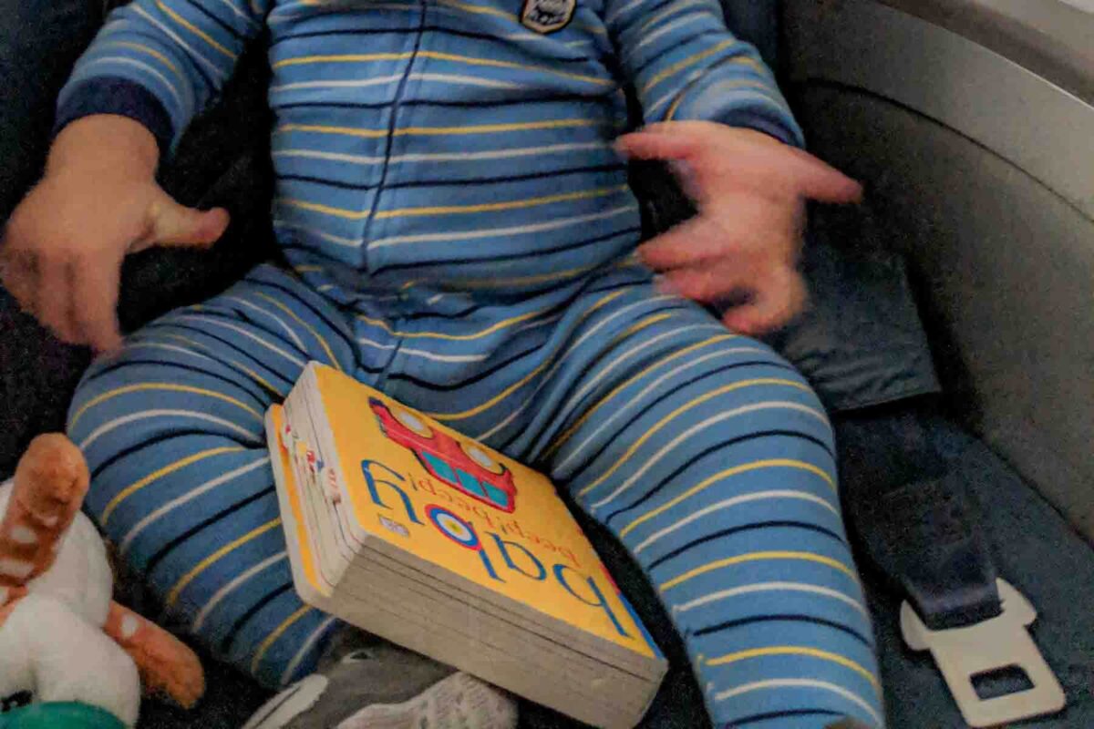 Baby in a striped blue pajama onesie sitting on an airplane seat with a small board book in their lap, showing how books work well as baby toys for airplane travel.
