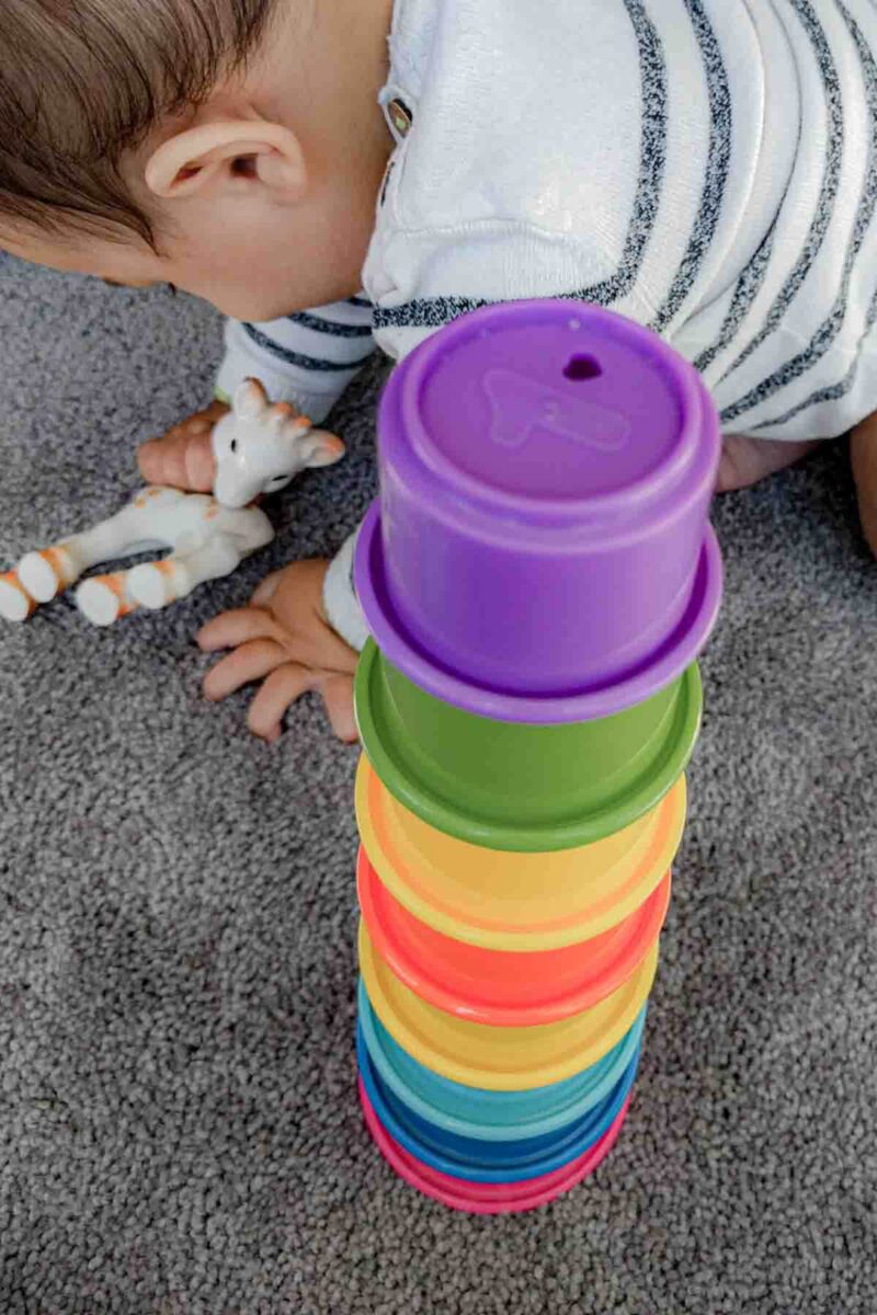 Baby playing with a tall stack of rainbow cups on a carpet while reaching forward, a classic baby travel toy that is lightweight and versitile.