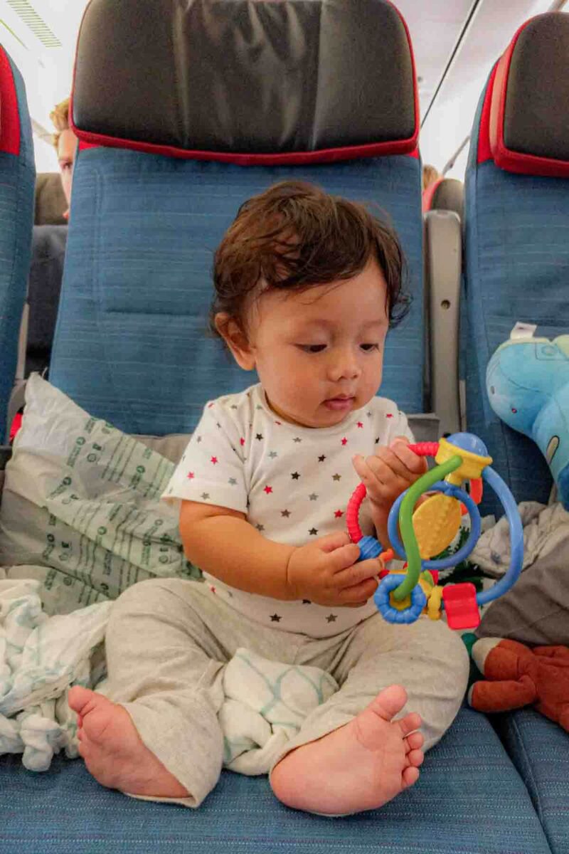 Six month old baby sitting on an airplane seat holding a colorful rattle toy, demonstrating how baby toys for airplane trips can keep little hands engaged.