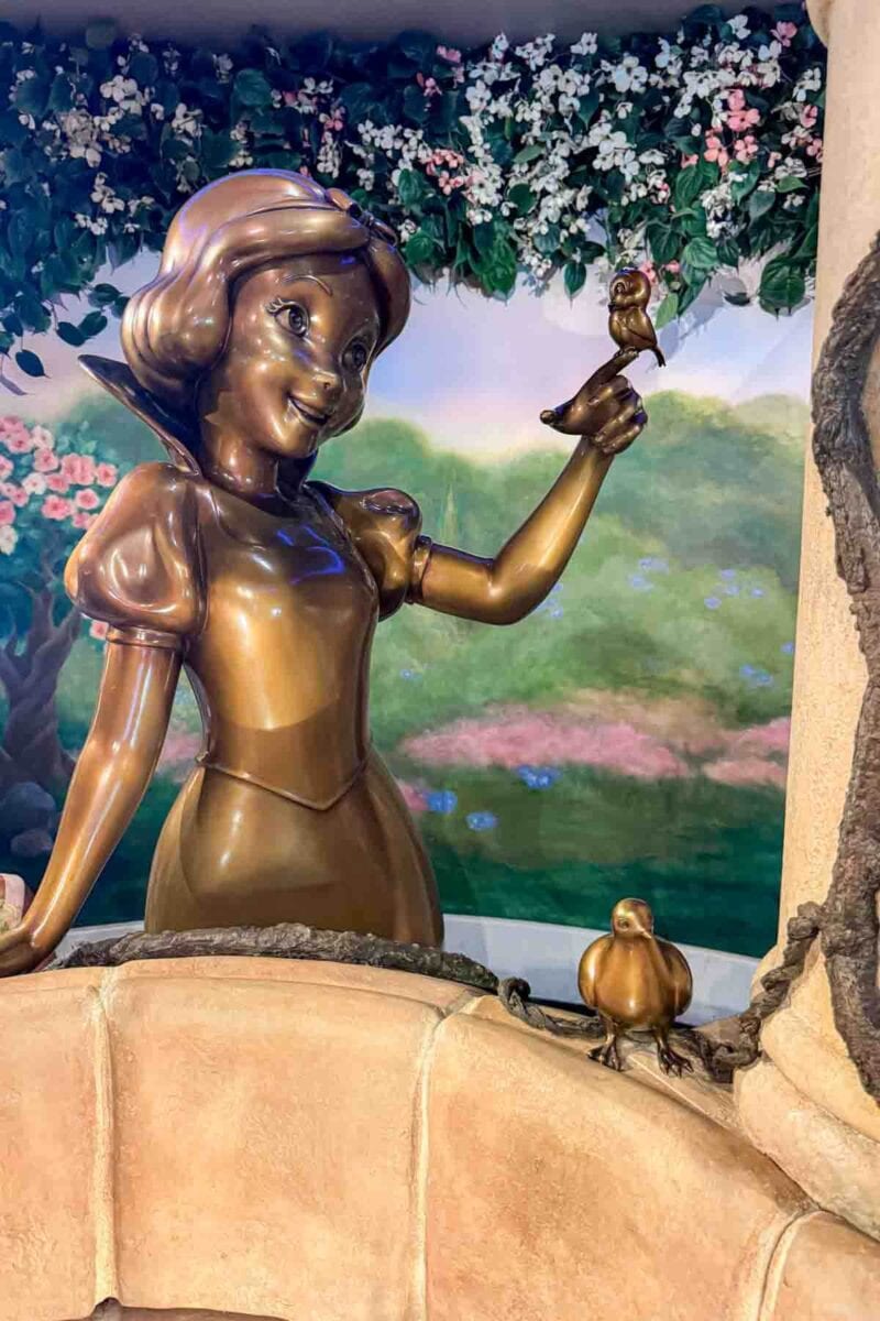 A bronze statue of Snow White smiling and holding a small bird stands beside a wishing well surrounded by floral details. This whimsical detail adds to the storytelling atmosphere in the Town Square onboard the Disney Adventure cruise ship.