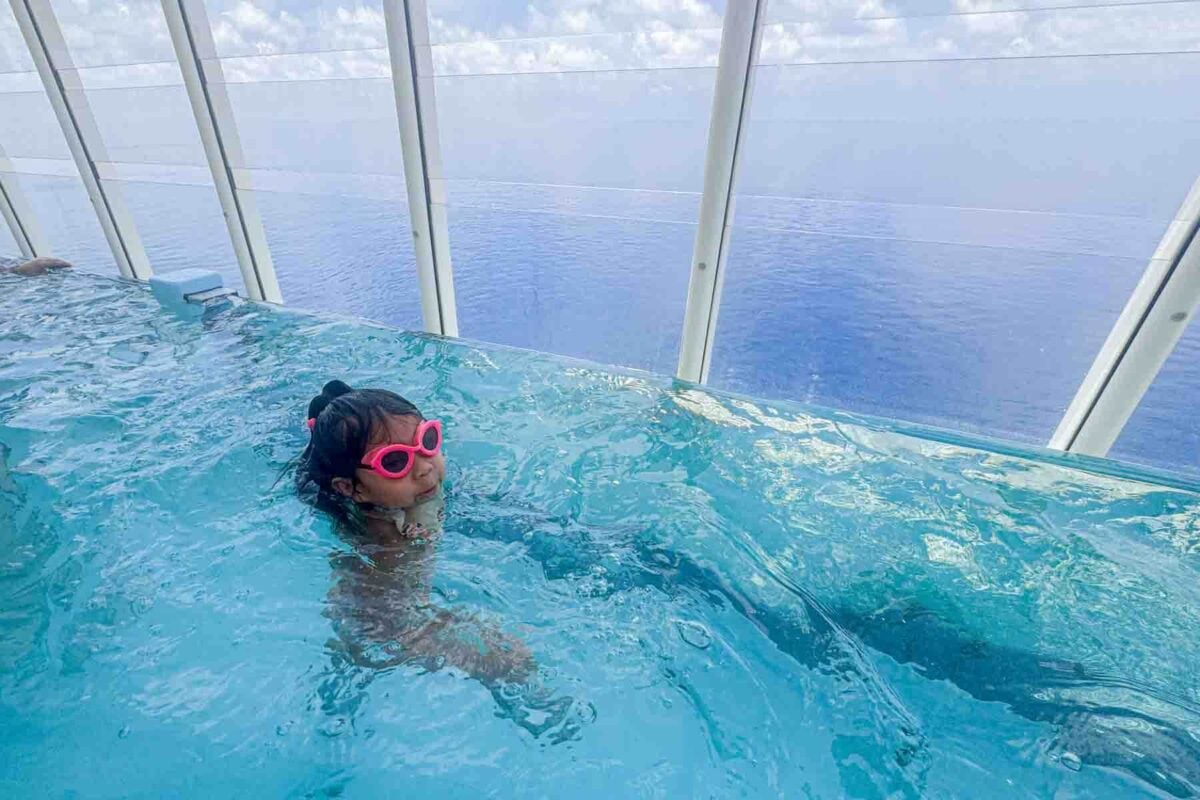 A child wearing pink goggles swims in an infinity style pool at the back of the Disney Adventure ship with open ocean views beyond glass panels. This scene highlights relaxing and fun Disney cruise activities for kids onboard.