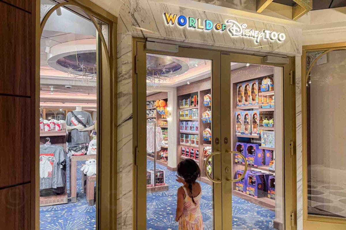 A child points excitedly toward the entrance of "World of Disney Too" shop filled with toys, clothing, and souvenirs. This moment showcases onboard shopping experiences for the Disney Adventure cruise.