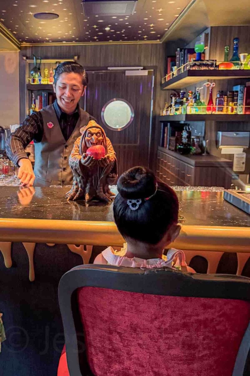 A child sits at a themed Spellbound lounge bar counter while a cast member smiles while making a drink. On the bar is a a detailed enchanted witch figure holding a glowing red apple. This interactive moment highlights themed experiences on the Disney Adventure cruise.