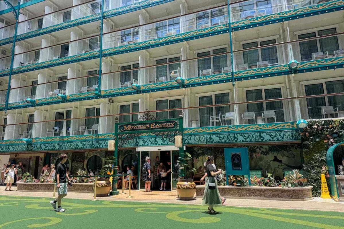 Guests walk through a lush courtyard space outside "Mowgli's Eatery" surrounded by greenery and interior verandah balcony staterooms above. This area showcases themed dining spaces and the design of Disney Adventure staterooms family layouts overlooking shared spaces.