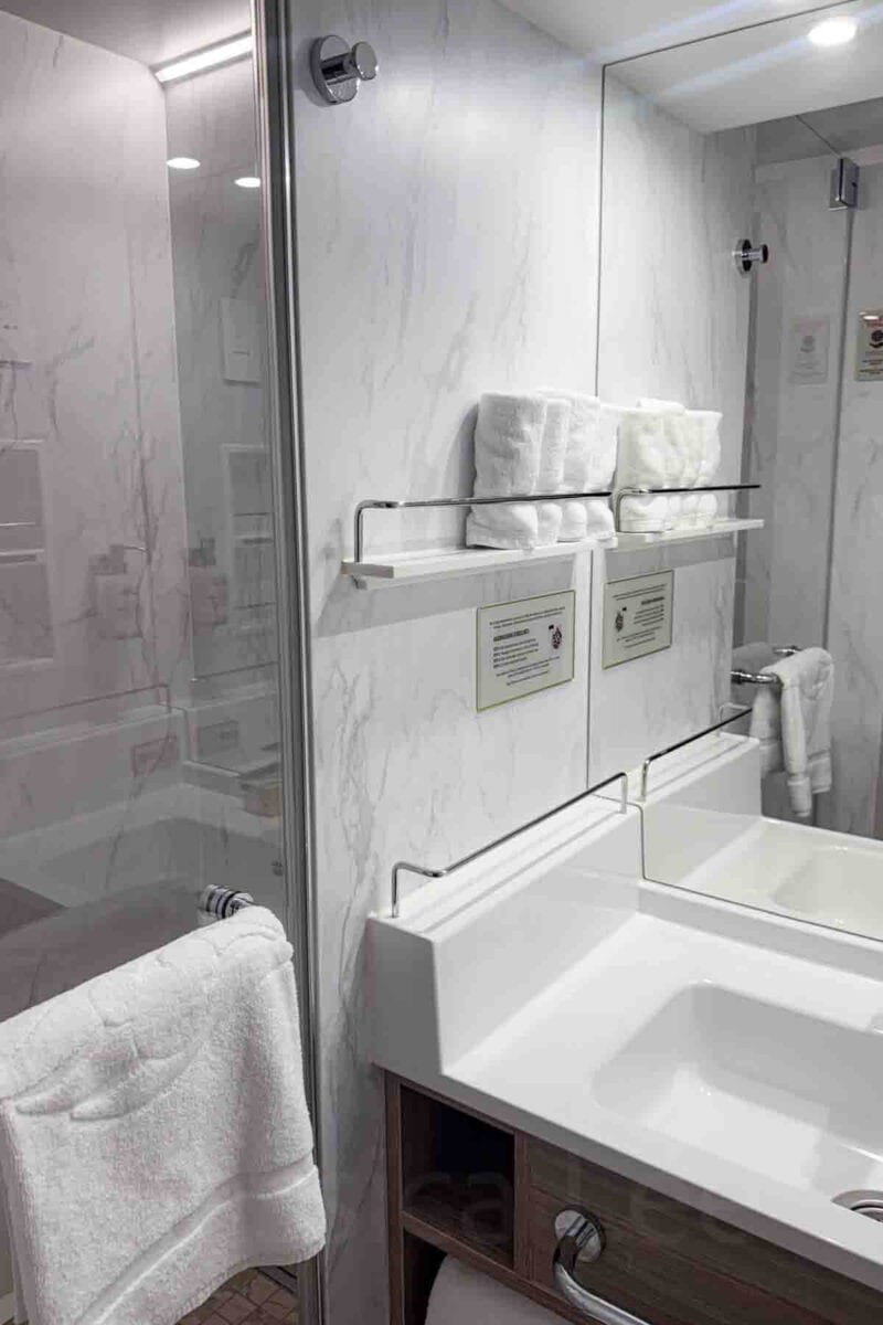 A clean cruise cabin bathroom features a white sink vanity, mirror, folded towels, and a glass enclosed shower. This setup demonstrates the layout of a Disney Adventure cruise interior stateroom bathroom.