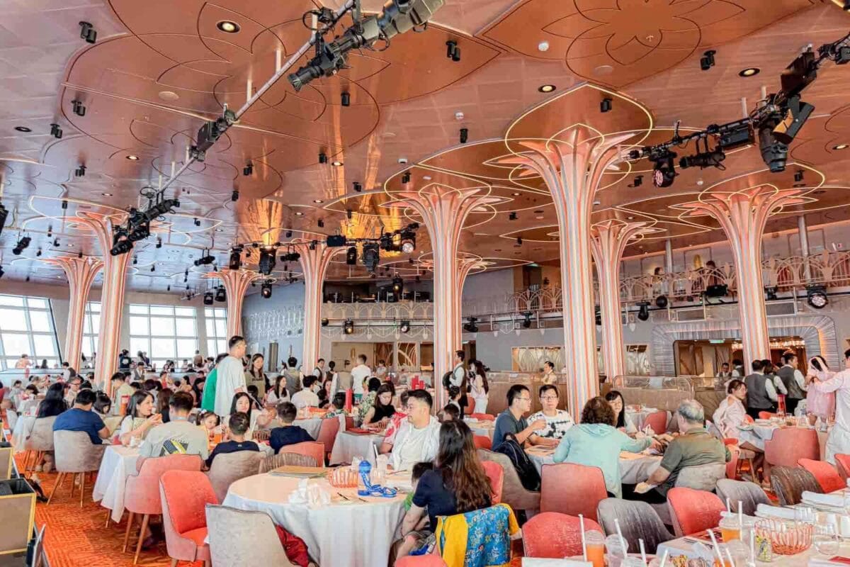 A lively expansive dining room filled with guests seated at round tables under glowing art deco style columns and warm lighting. This scene shows the energetic atmosphere of dinner at the Hollywood Spotlight Club on the Disney Adventure cruise.