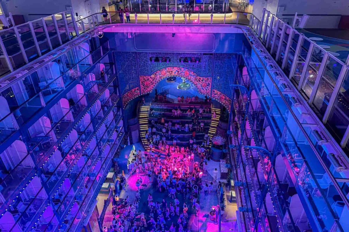 An overhead view of a multi level atrium shows a large crowd gathered around a central stage during a nighttime dance party with purple and blue lighting. This highlights evening entertainment in the Imagination Garden on Disney Adventure cruise.