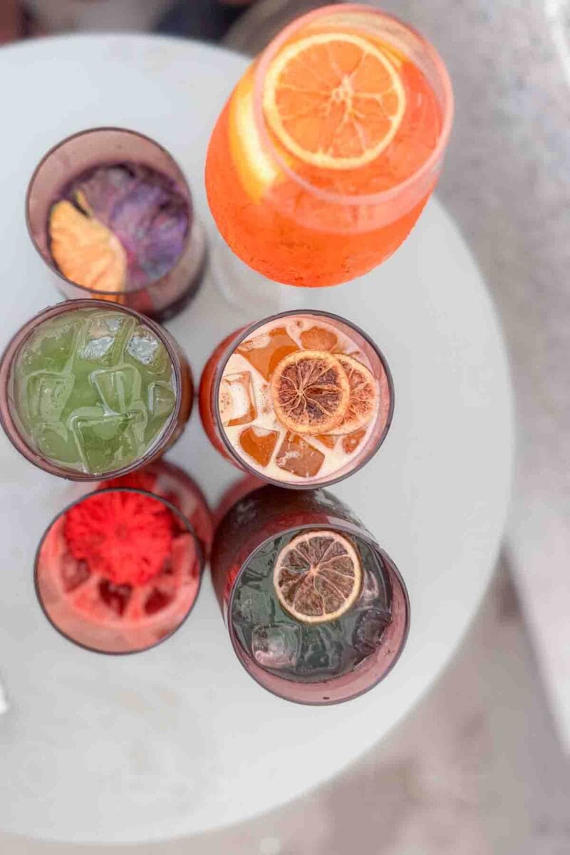 A top down view of colorful cocktails garnished with citrus slices and ice sits on a round table. This image showcases specialty “Infitinity Stone” Marvel themed drinks served at a Disney cruise bar experience.