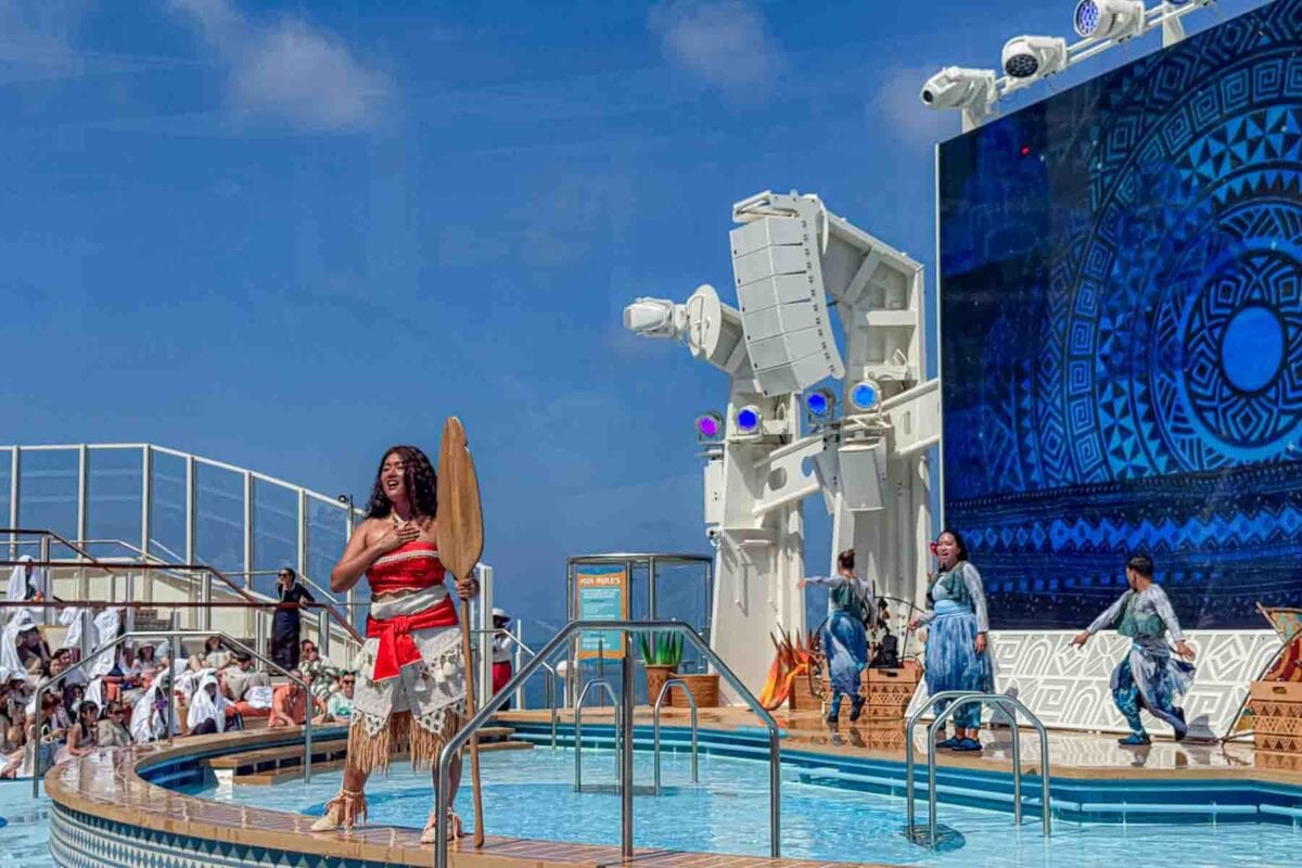 A performer dressed as Moana stands while singing by a small pool holding an oar while dancers perform behind her on an outdoor stage. This live show highlights family entertainment on the Disney Adventure cruise.