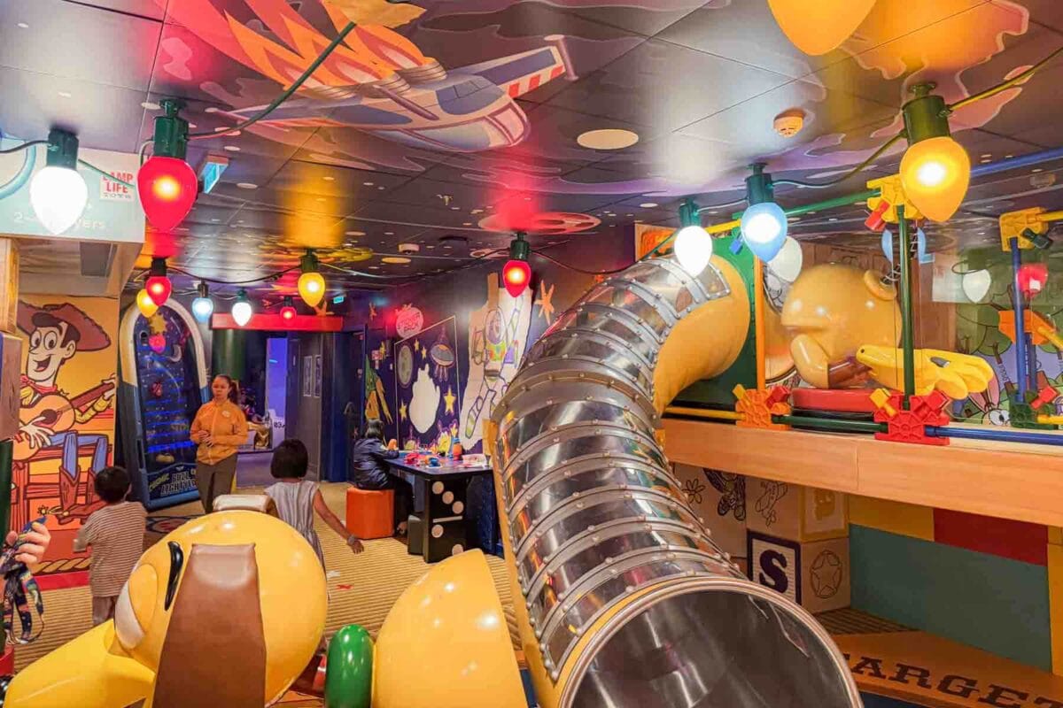 A colorful Toy Story themed play area features a tube slide, oversized character decor, and hanging lights shaped like toys. This space showcases imaginative play zones inside the Disney Adventure cruise kids club.