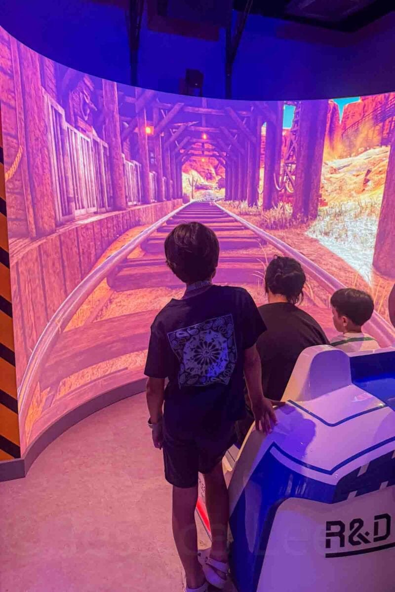 Children sit in a ride vehicle facing a curved screen showing a roller coaster track through a canyon landscape. This interactive simulator highlights fun experiences for kids inside the Oceaneer Club on the Disney Adventure cruise.