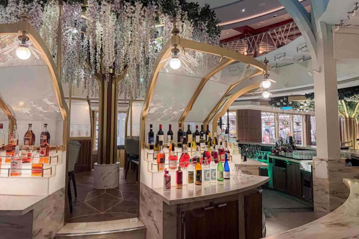A stylish round bar displays rows of colorful liquor bottles beneath hanging greenery and warm lighting. This setting highlights the elegant atmosphere of Tiana’s Bayou lounge on Disney Adventure cruise.