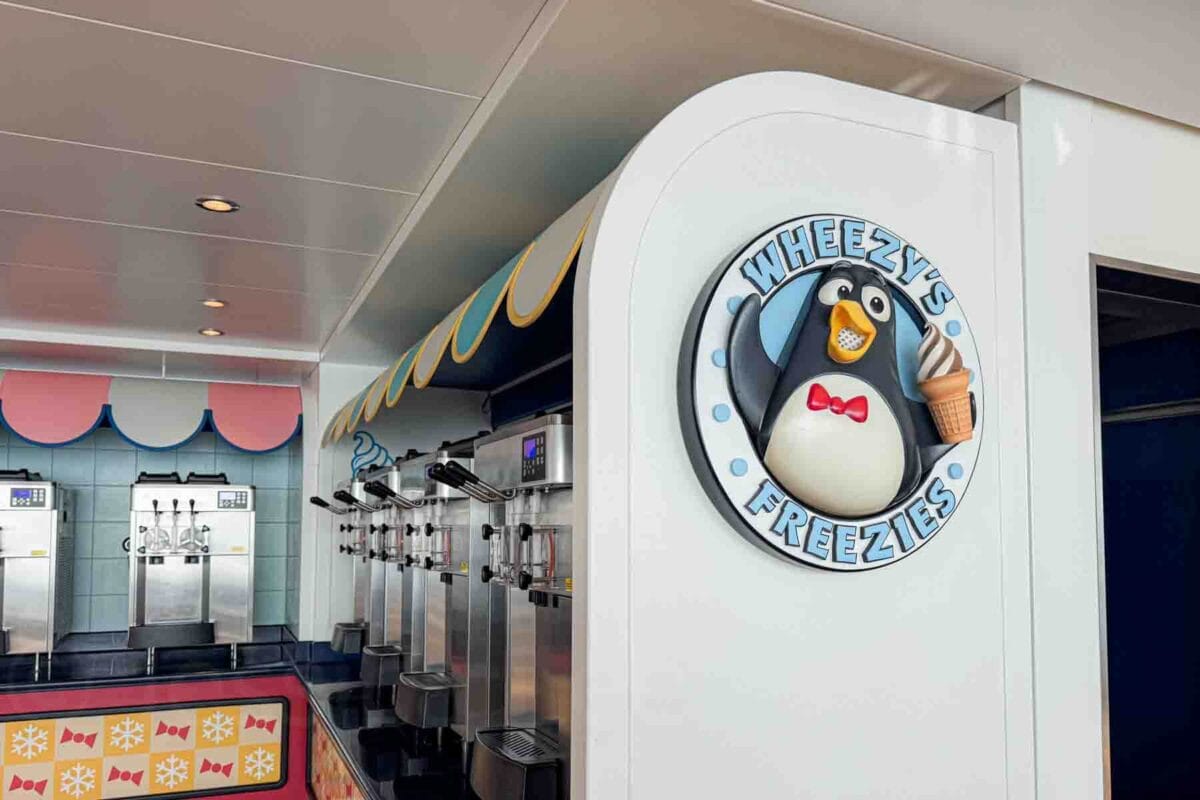 A soft serve ice cream station with multiple machines sits beside a sign reading "WHEEZY'S FREEZIES" featuring a cartoon penguin holding an ice cream cone. This spot highlights a fun, casual dessert option kids love on the Disney Adventure cruise.
