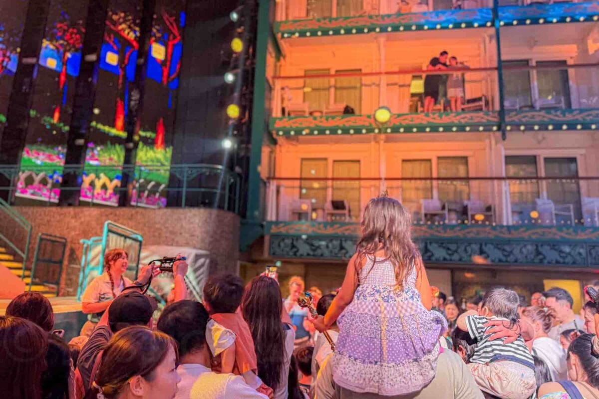 Families gather in a central courtyard, the Imagination Garden, with bright screens and festive lighting dancing with classic Disney characters. This scene shows how entertainment brings crowds together during a Disney Adventure cruise.