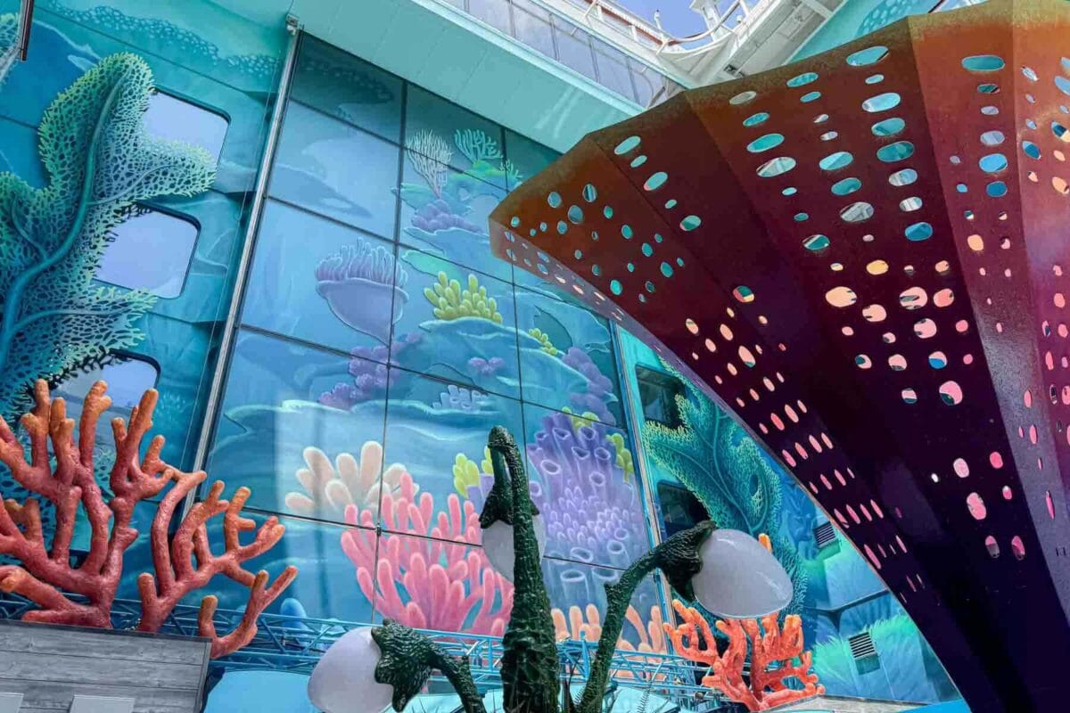 A vibrant underwater themed area features coral shaped sculptures and a mural of ocean life in shades of blue, pink, and green. This immersive design showcases the Discovery Reef themed zone on the Disney Adventure cruise.