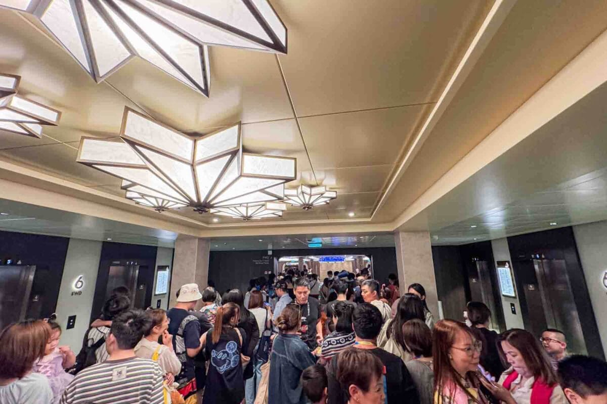 A large crowd of guests lines up in a hallway near elevators under decorative ceiling lights awaiting a show. This moment highlights peak time crowds on a Disney Adventure cruise.
