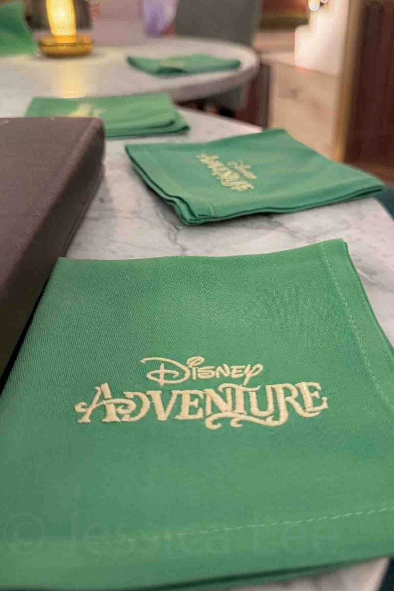 A neatly folded green napkin embroidered with "Disney Adventure" sits on a dining table with soft lighting. This detail highlights branded touches included in a Disney cruise experience.