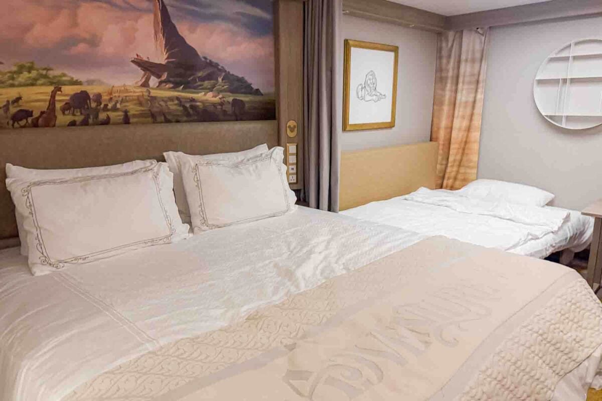 A cozy interior stateroom shows a large bed and convertible sofa bed with a Lion King themed artwork above the headboard. This room setup highlights family friendly Disney Adventure cruise stateroom design.