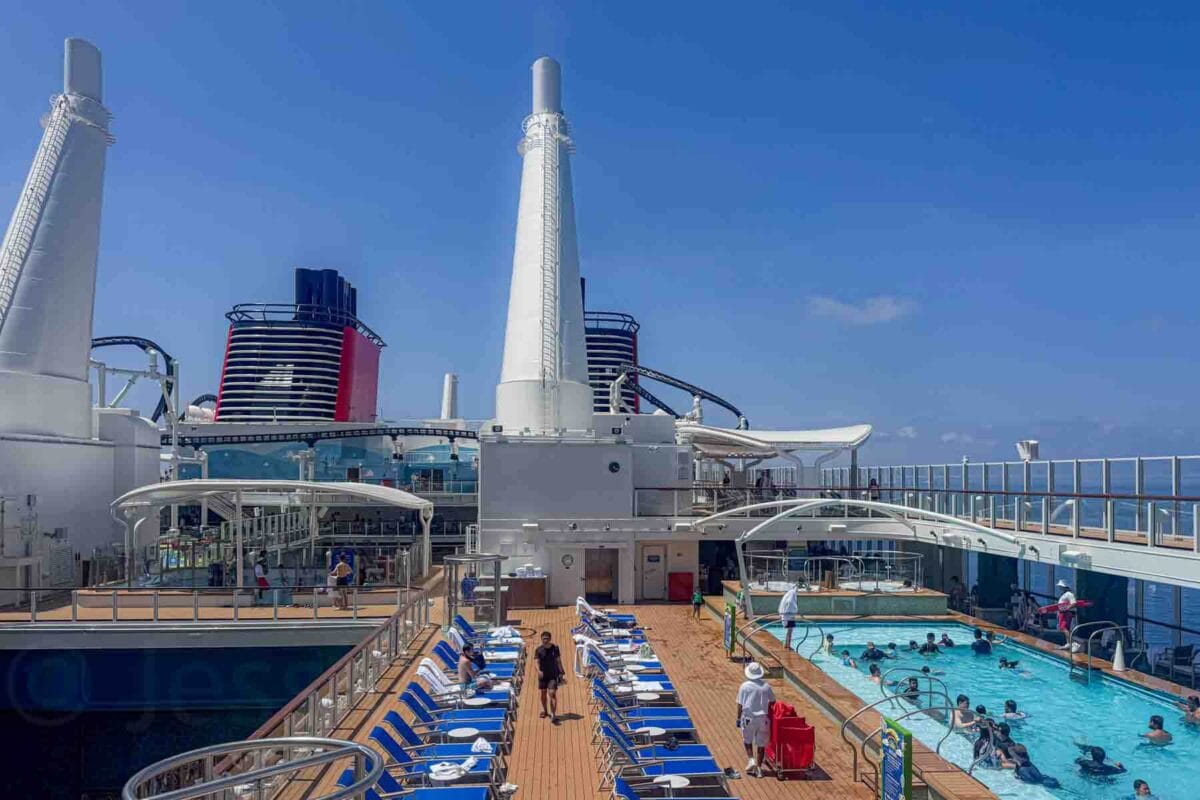 Rows of blue lounge chairs line the pool deck 17 beside a swimming pool with guests relaxing and walking nearby. This scene shows the Disney Adventure cruise pool deck seating and relaxation areas.