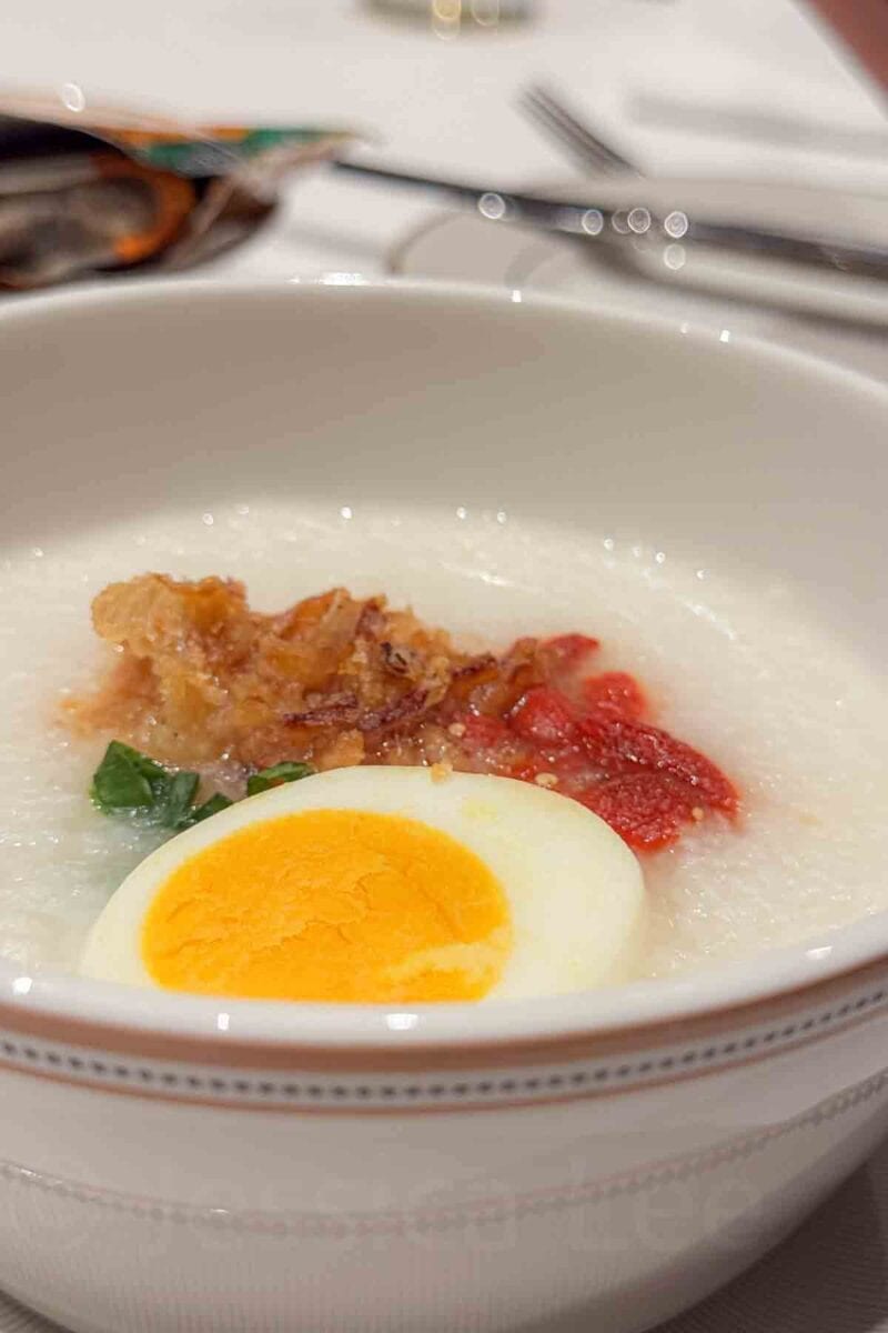 A bowl of congee topped with a soft boiled egg, crispy toppings, and greens sits on a dining table. This dish reflects breakfast food options included in a Disney Adventure cruise.