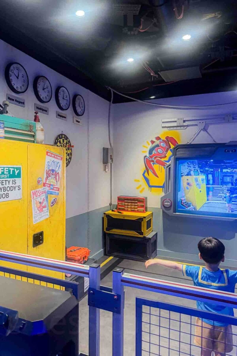 A child stands inside a Marvel themed play space with screens, tools, and wall graphics including a red spider emblem. Visible text includes "SAFETY FIRST SAFETY IS EVERYBODY'S JOB" on a cabinet. This area highlights themed interactive kids activities in the Oceaneer Club on Disney Adventure cruise.