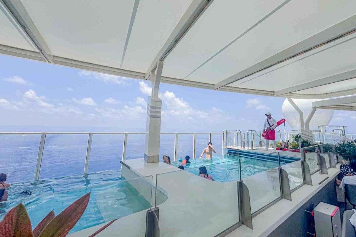 Guests relax in an infinity pool overlooking the ocean with glass railings, decorative plants, and a shaded deck area. This pool area shows one of the scenic relaxation spots on Disney Adventure cruise.