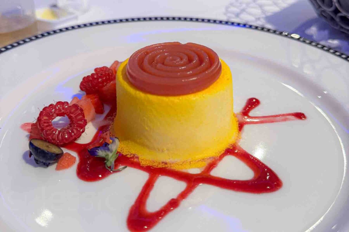 A plated mango dessert topped with a spiral of red sauce is surrounded by fresh raspberries, flowers, and fruit garnish. This dish highlights elegant dessert offerings on a Disney cruise dining menu.