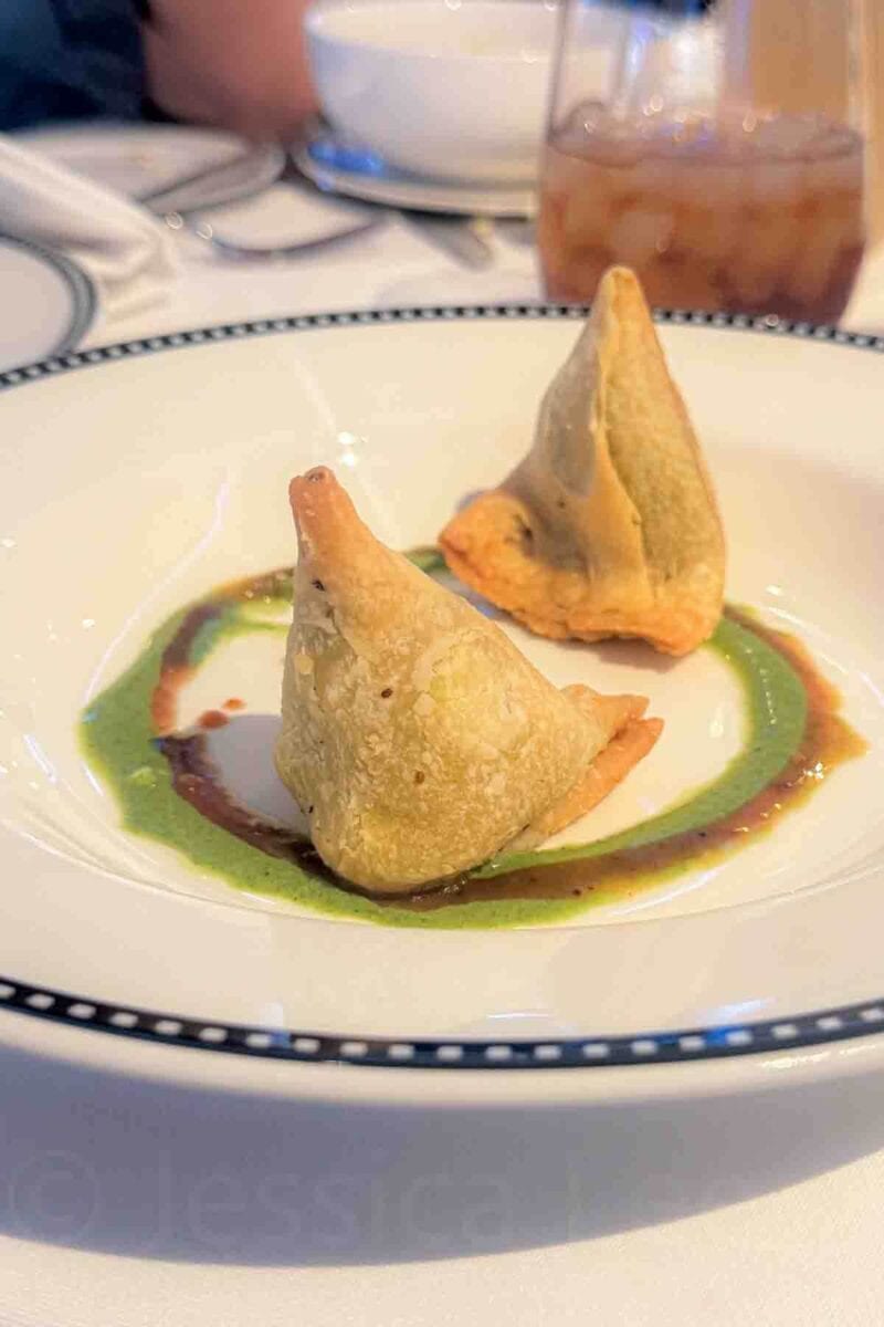Two golden fried samosas are plated with green and brown sauces arranged in a circle underneath. This appetizer showcases international options served during a Disney cruise.