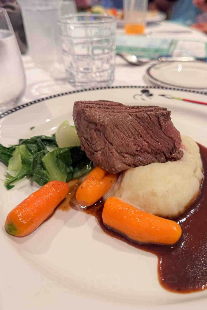 A thick cut of steak sits on mashed potatoes with baby carrots and greens, topped with a rich brown sauce. This entrée highlights classic fine dining options on a Disney cruise.