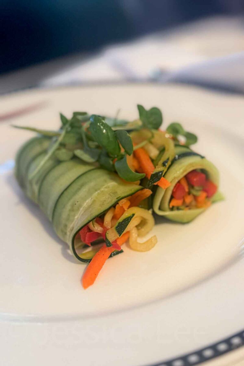 Thinly sliced cucumber wraps around colorful vegetables and is topped with microgreens on a white plate. This dish showcases a fresh and light appetizer option on a Disney cruise.