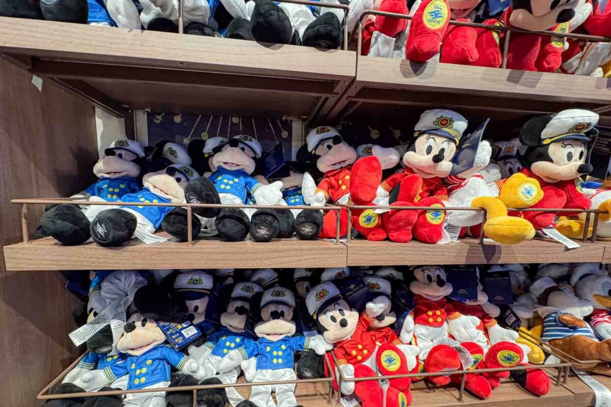 Shelves filled with Mickey and Minnie plush toys dressed in cruise uniforms are displayed in a shop. This merchandise area showcases souvenirs available onboard a Disney cruise.