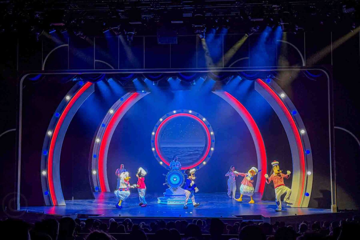 Mickey, Daisy, Donald, and Goofy perform on a brightly lit stage with colorful circular set designs and ocean backdrop. This live show highlights onboard entertainment on a Disney cruise.