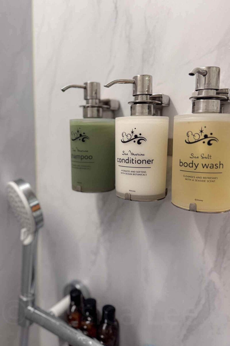 Wall mounted bottles labeled "shampoo", "conditioner", and "body wash" are arranged neatly in a cruise ship shower. These amenities highlight in room bathroom features on a Disney cruise.