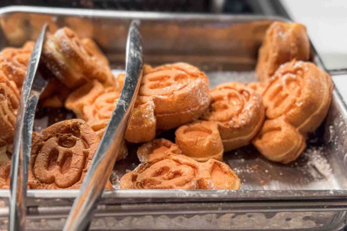 Golden waffles shaped like Mickey Mouse sit in a tray with serving tongs and powdered sugar. This breakfast item highlights a classic Disney cruise dining favorite among kids.
